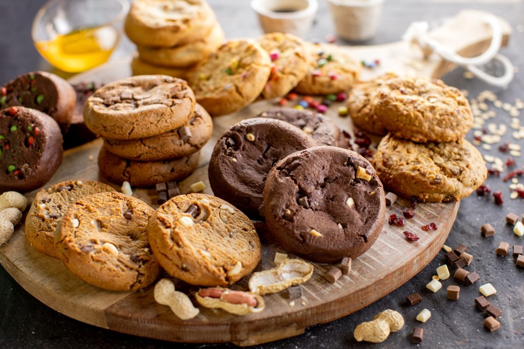 De Graaf Bakeries | product categorie | American cookies | banner waarop je op een borrelplank verschillende soorten cookies te zien krijgt. Om de koeken heen liggen ingredienten die in de koeken zijn gebruikt zoals stukjes chocolade, smarties en pinda's.